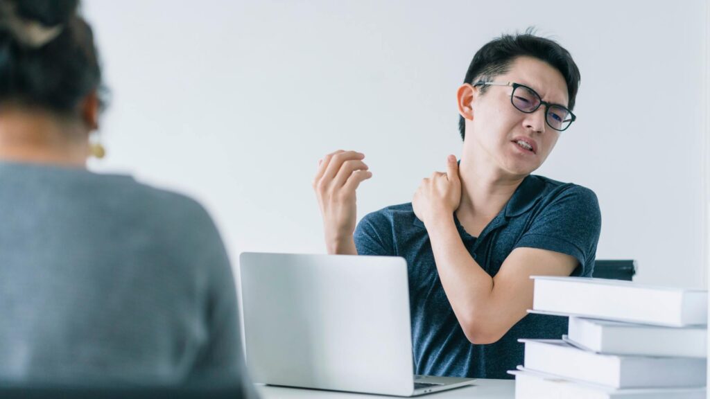 Pria pekerja kantoran memegang bahu yang terasa nyeri dan pegal akibat terlalu lama bekerja di depan laptop, menggambarkan kondisi badan pegal terus karena aktivitas dan postur kerja yang kurang ideal.
