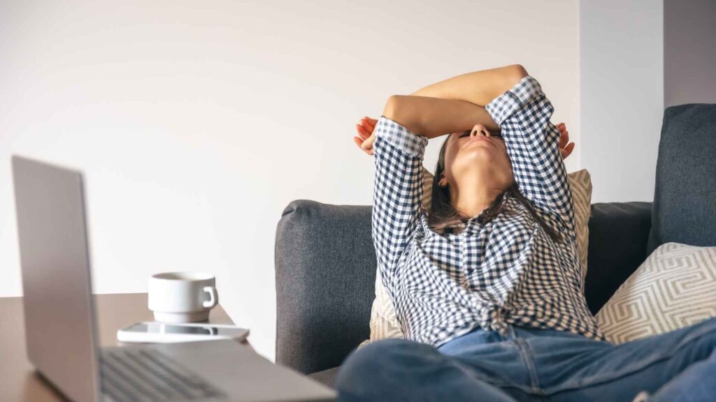 Wanita bersandar di sofa sambil menutupi wajah dengan tangan di depan laptop sebagai tanda tubuh butuh istirahat setelah bekerja seharian.